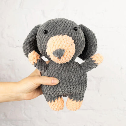 A hand is holding a gray and peach-colored stuffed animal, which appears to be a small dog or puppy.