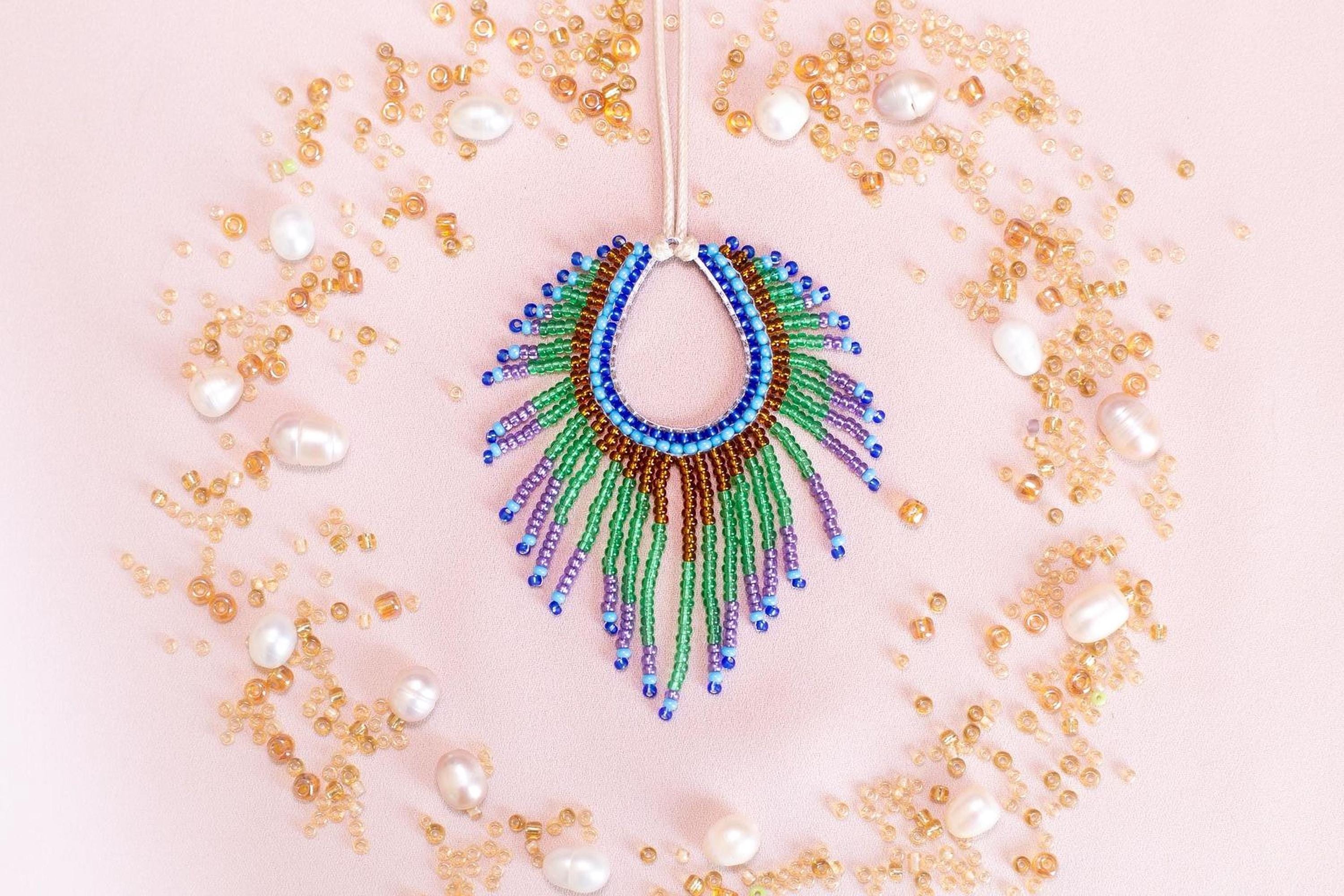 a colorful beaded necklace with a large, eye-catching pendant surrounded by a variety of small, round, and multicolored beads, pearls, and other decorative elements arranged in a circular pattern on a pink background.