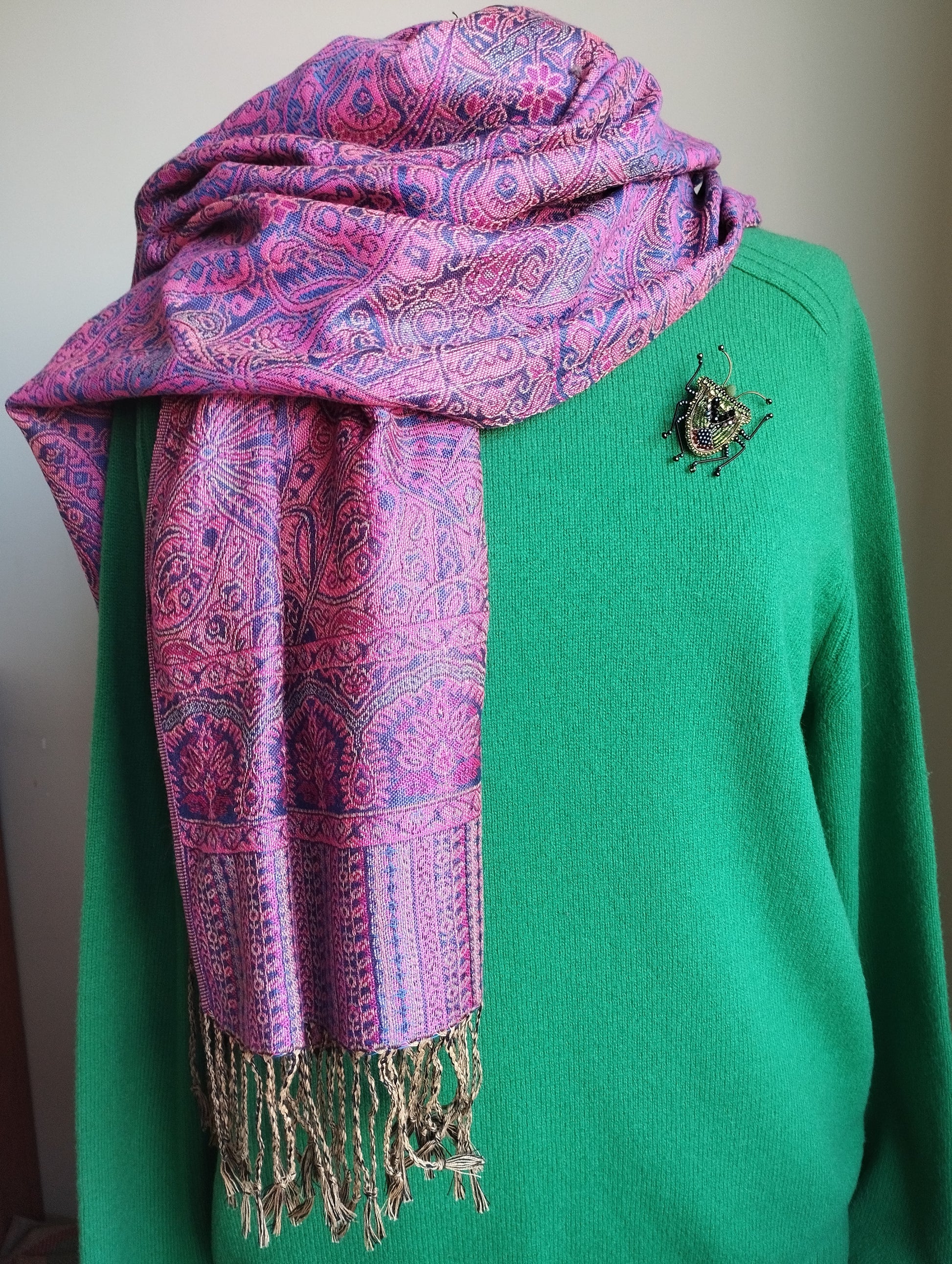 A mannequin is wearing a green sweater and a pink and purple paisley-patterned scarf with fringe. The scarf is draped over the sweater, creating a stylish and coordinated outfit.