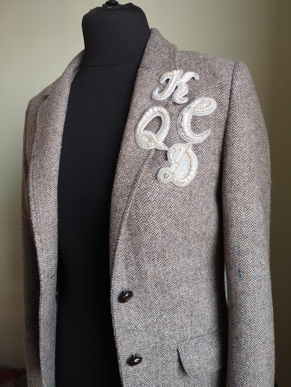 A gray tweed jacket with a white embroidered letter on the left side of the chest.