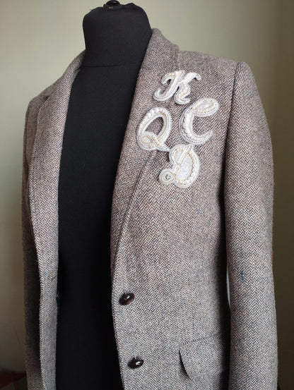 A gray tweed jacket with a white embroidered letter on the left side of the chest.