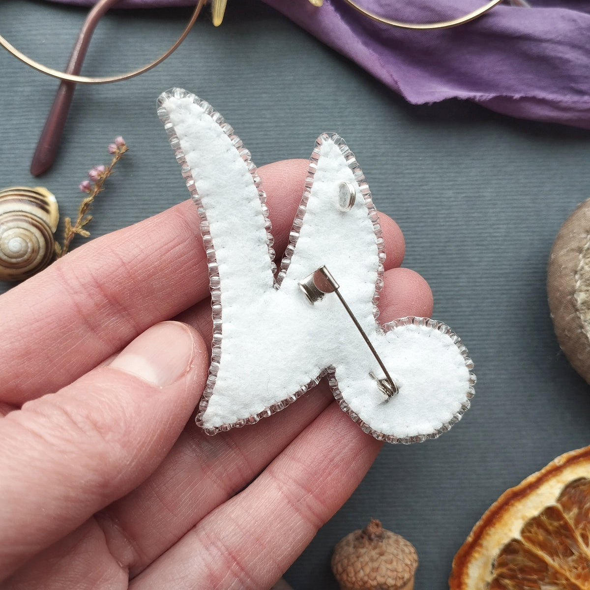A hand holding a white felt letter "V" with a pin attached to it, surrounded by various objects such as a pair of glasses, a purple fabric, and dried fruit.