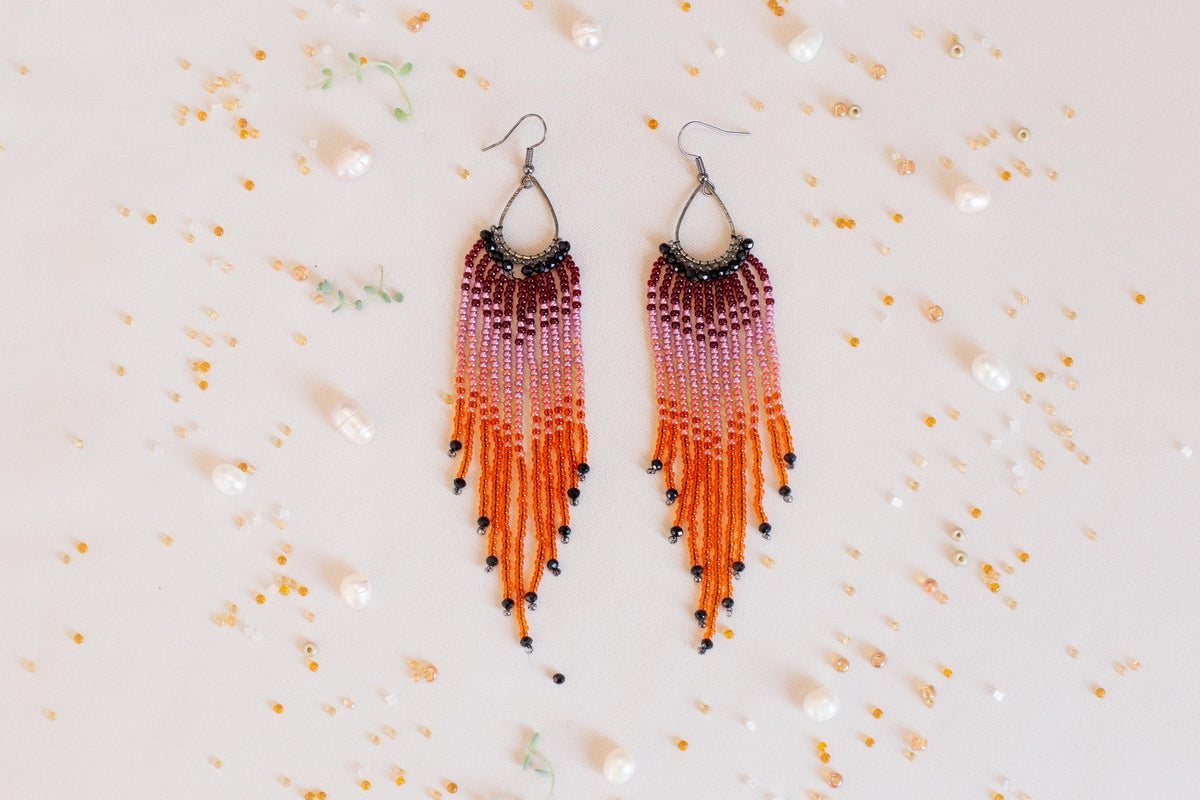 a pair of earrings with long, dangling strands of beads in shades of orange and purple, arranged on a white background. The earrings are surrounded by scattered beads and pearls, creating a visually appealing and colorful composition.