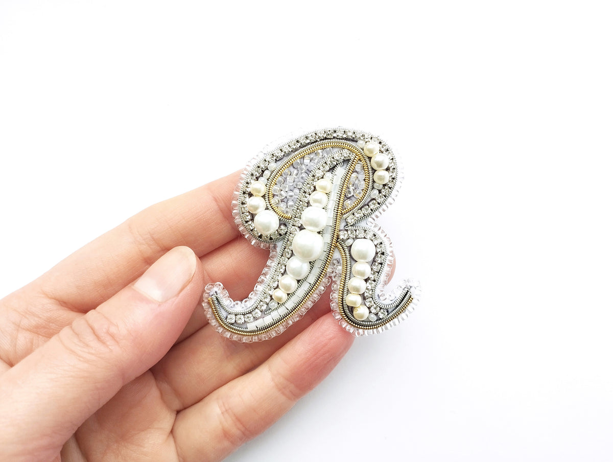 A hand holding a decorative letter "R" made of pearls and rhinestones against a white background.