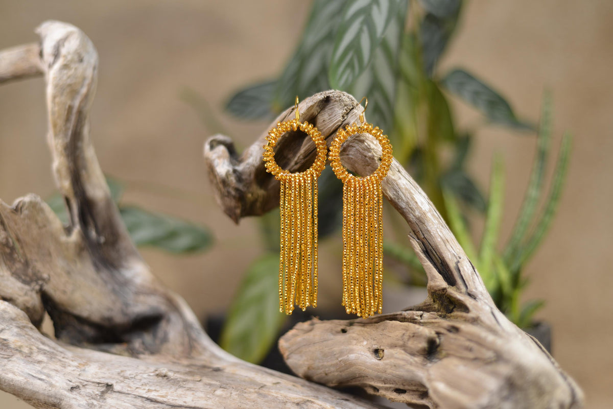 A pair of gold earrings with dangling tassels, displayed on a wooden branch against a blurred background of plants.