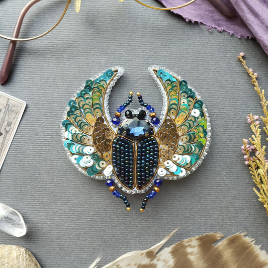 a detailed embroidered piece of jewelry, resembling a butterfly or a beetle, with intricate patterns and designs. The piece is adorned with various beads and sequins, giving it a vibrant and eye-catching appearance. The jewelry is displayed on a gray surface, surrounded by other objects such as feathers and crystals, creating a visually appealing and artistic composition.