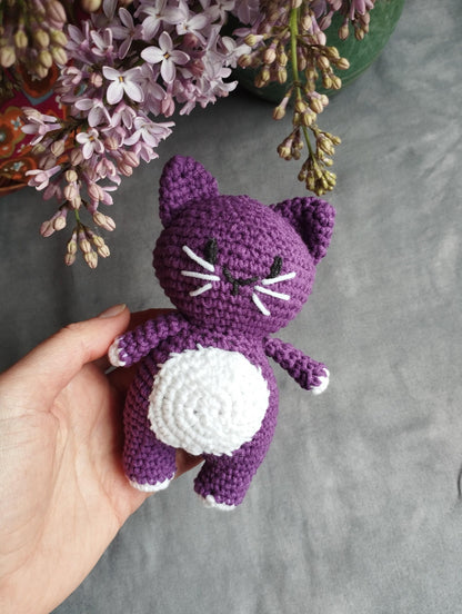 a hand holding a purple crocheted cat next to a bouquet of flowers