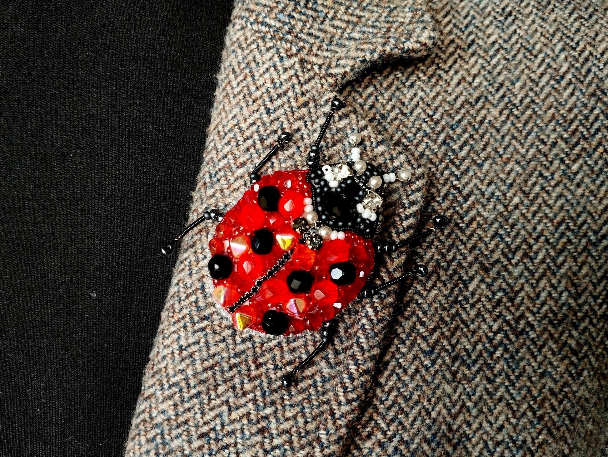A close-up image of a red and black ladybug brooch with beaded accents, attached to a grey tweed jacket.