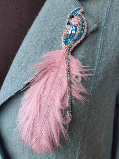 A close-up image of a light blue fabric with a pink feather attached to it, featuring a decorative brooch-like embellishment.
