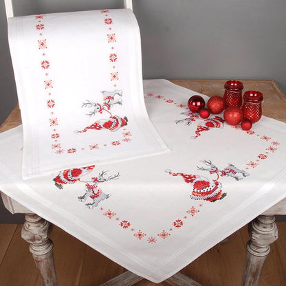 a white tablecloth with a red and white Christmas-themed design, including reindeer and snowflakes. The tablecloth is placed on a wooden table, and there are red ornaments and candles nearby.