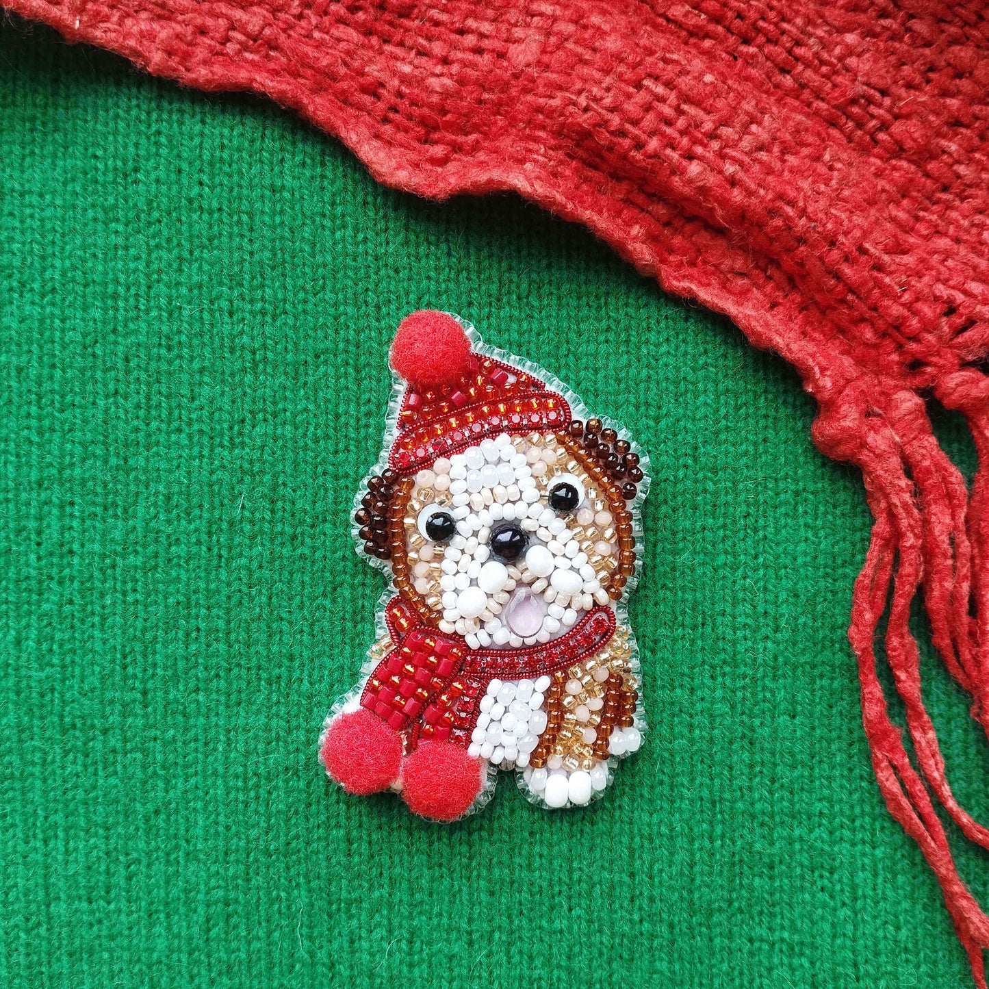 A small embroidered dog wearing a red hat and scarf is displayed against a green background, with a red scarf partially visible on the right side.