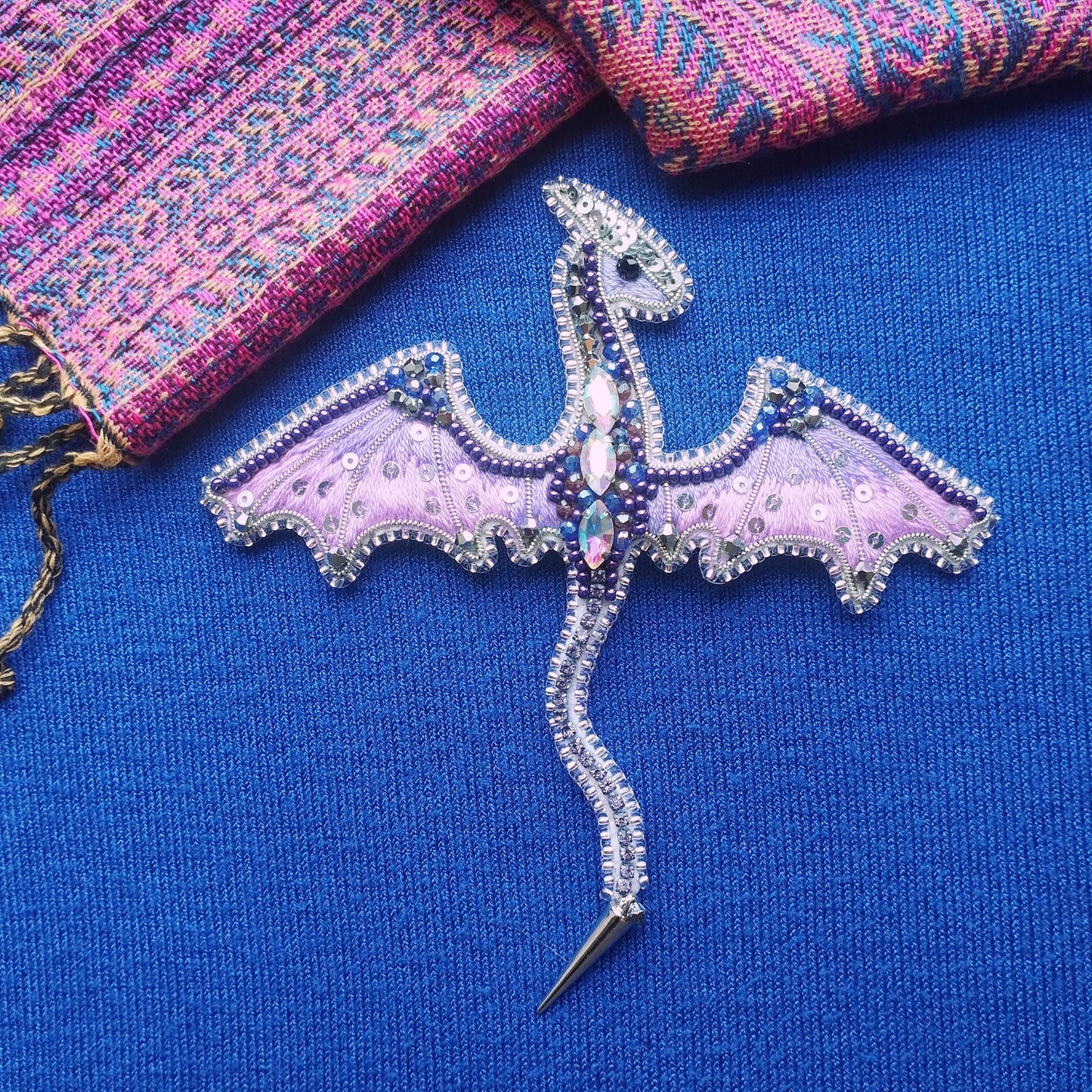 A decorative dragon pin with intricate beading and gemstone details rests on a blue fabric surface.