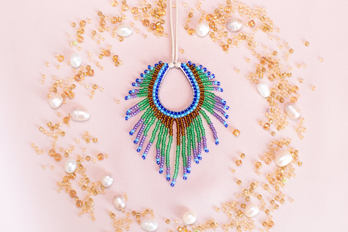 a colorful beaded necklace with a large, eye-catching pendant surrounded by a variety of small, round, and multicolored beads, pearls, and other decorative elements arranged in a circular pattern on a pink background.