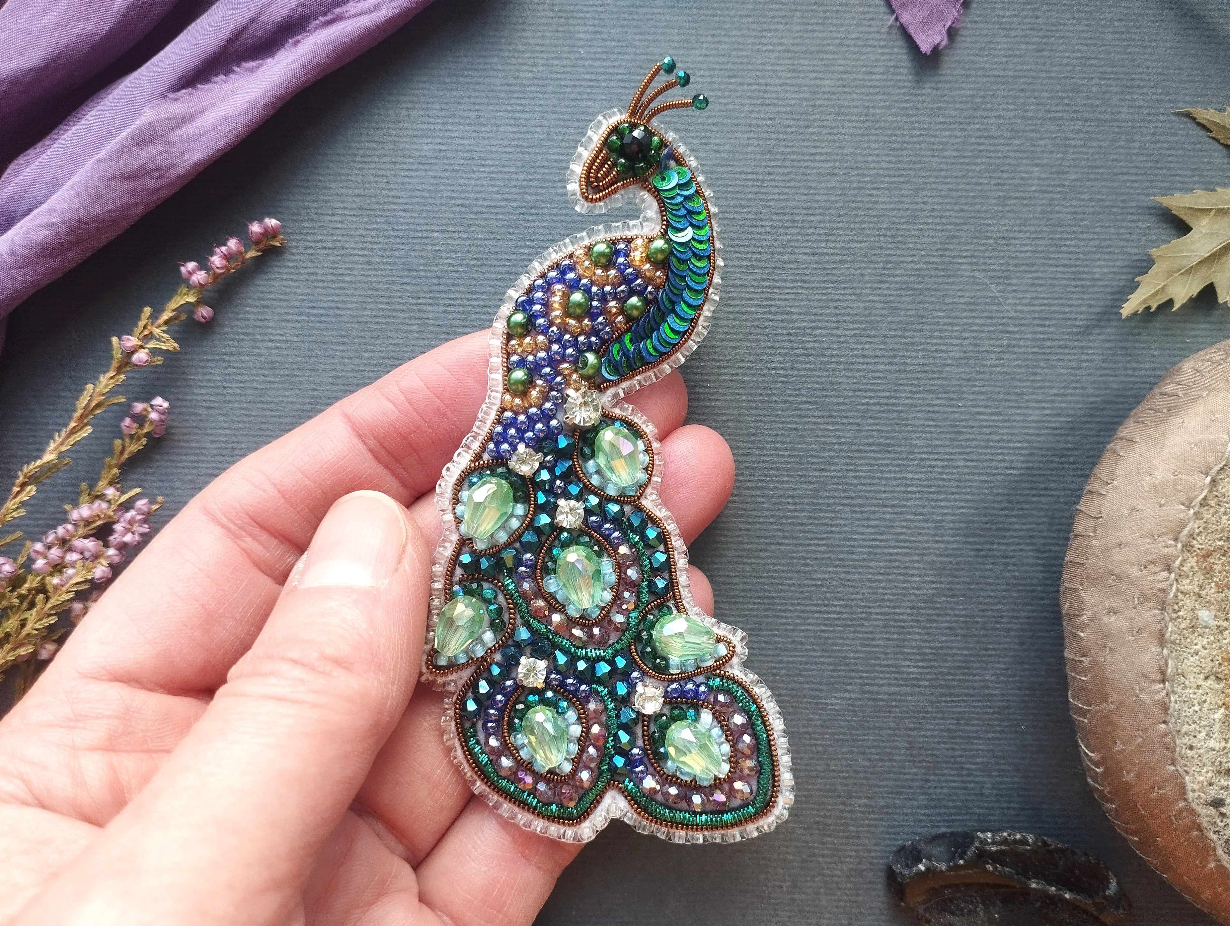 A hand holding a decorative peacock brooch with colorful beads and stones.