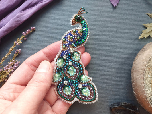 A hand holding a decorative peacock brooch with colorful beads and stones.