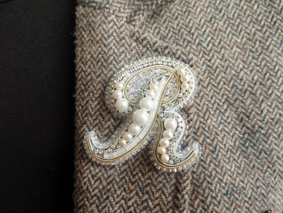 A close-up image of a decorative monogram made of pearls and beads, with a dark fabric background.