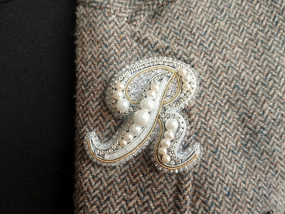 A close-up image of a decorative monogram made of pearls and beads, with a dark fabric background.