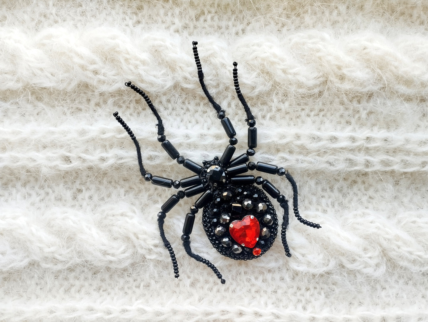 A black spider-shaped brooch with red gemstones is displayed on a white, knitted fabric.