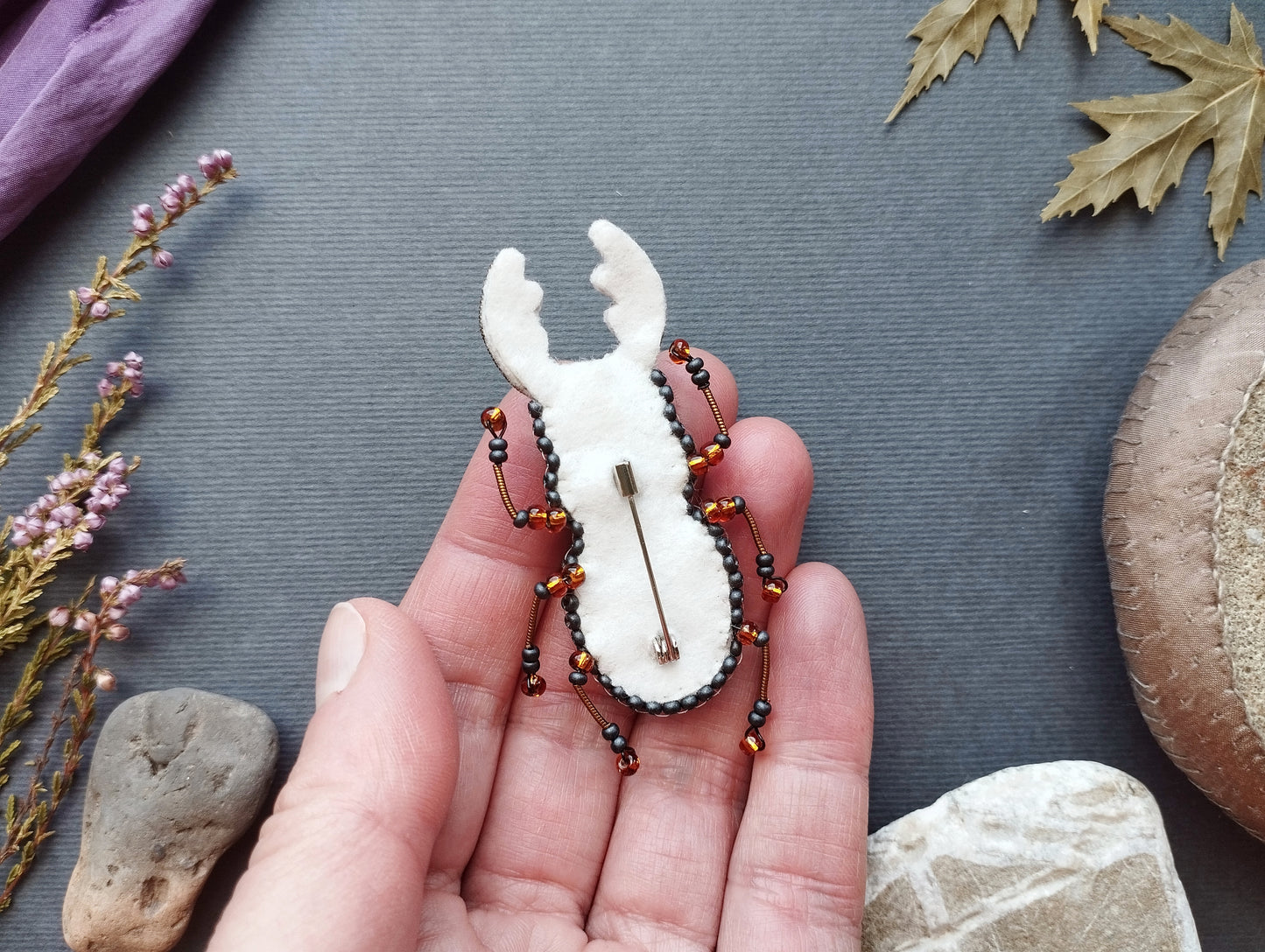 A hand holding a white, beaded insect brooch against a gray background with various natural elements, such as leaves and rocks.