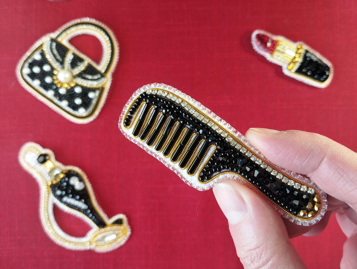 A hand is holding a black comb with gold and black beads, against a red background with various decorative elements, including a handbag and a lipstick.