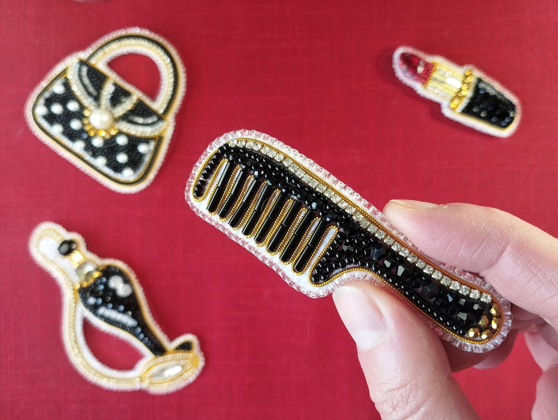 A hand is holding a black comb with gold and black beads, against a red background with various decorative elements, including a handbag and a lipstick.