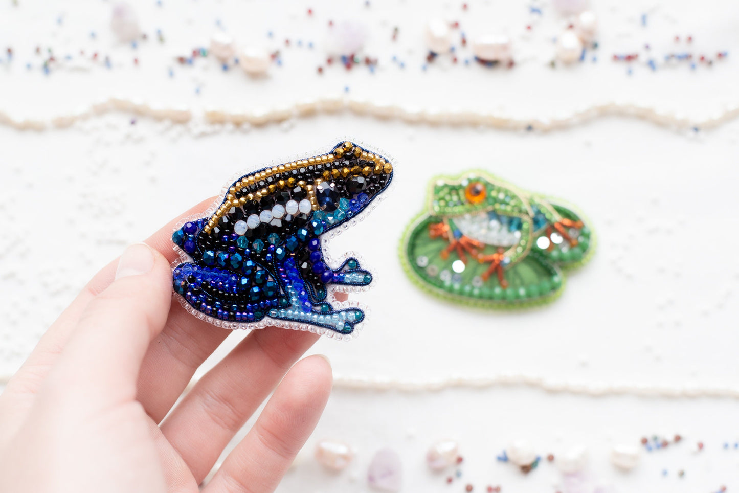 a hand holding a beaded frog next to another beaded frog