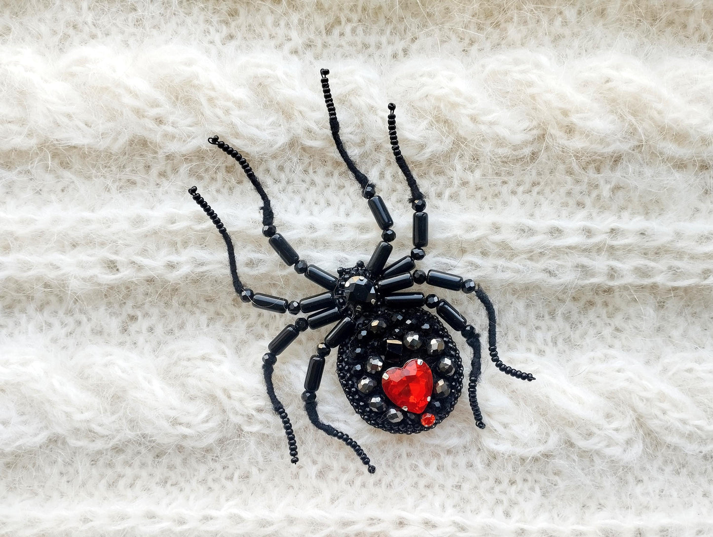 A black spider-shaped brooch with red gemstones is displayed on a white, knitted fabric.