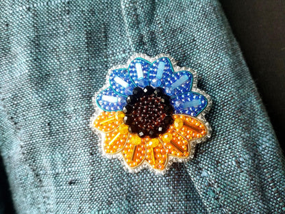 A close-up view of a colorful, intricately designed pin or brooch featuring a sunflower motif, with a mix of blue, orange, and brown beads and stones.