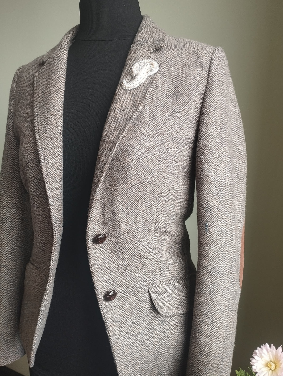 A gray tweed blazer with a white flower pinned to the lapel, displayed on a mannequin.