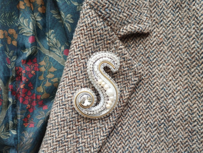 A close-up image of a decorative letter 'S' brooch on a tweed jacket, with a floral patterned fabric visible in the background.