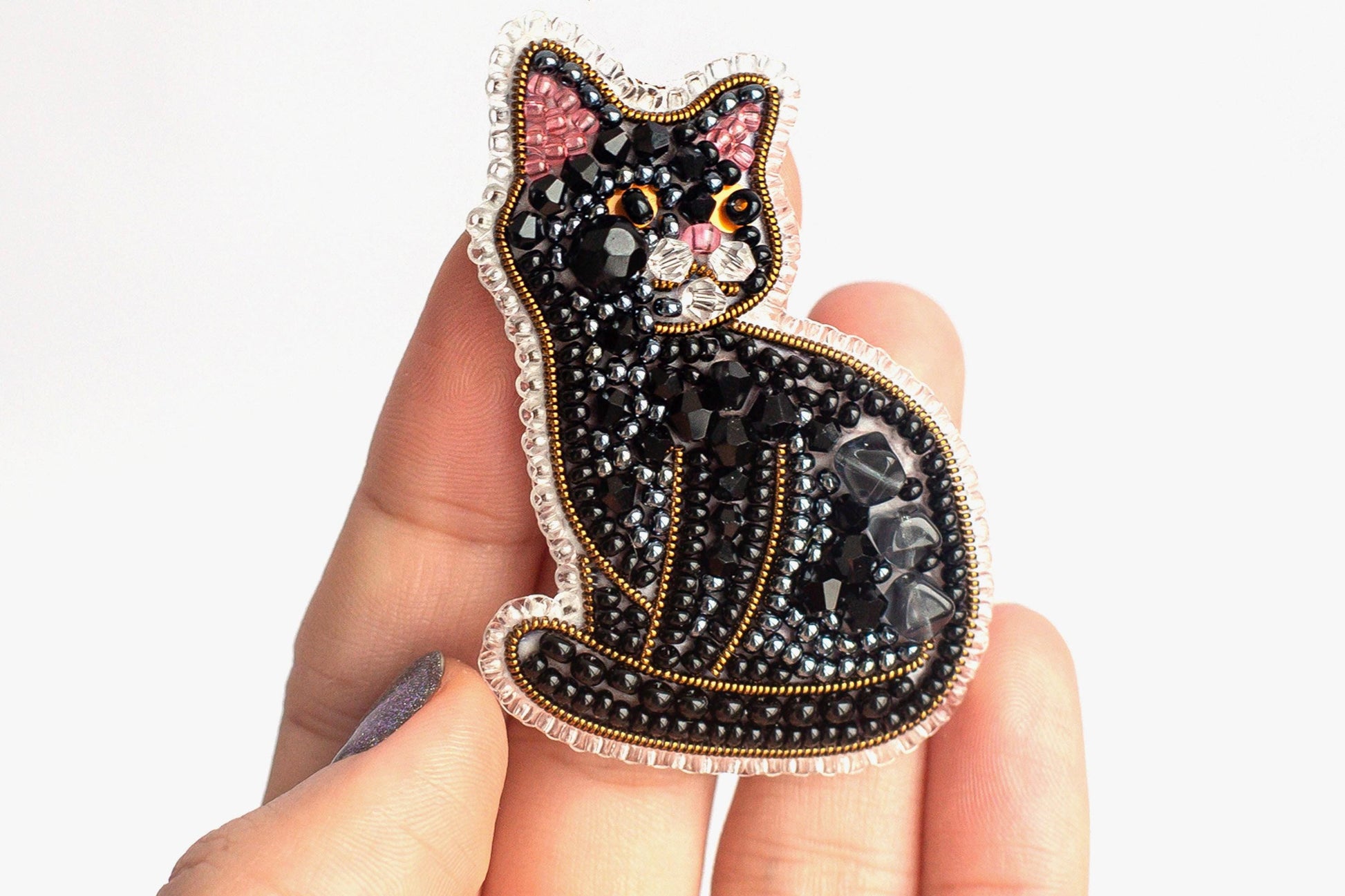 A hand holding a decorative cat pin made of black beads and gold accents.