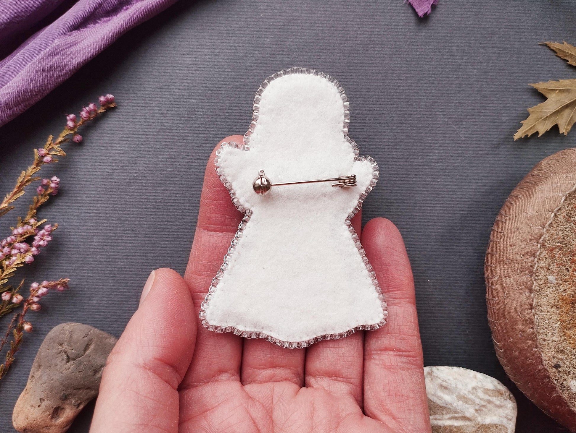 A hand holding a white felt ghost pin against a dark background with various objects, including a leaf, a rock, and some flowers.