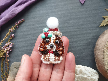 A hand holding a small, intricately beaded brooch depicting a dog wearing a hat and scarf.