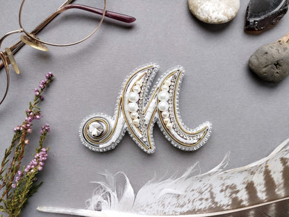 a close-up view of a decorative letter "M" made of beads, pearls, and feathers, surrounded by various accessories such as glasses, stones, and a feather.
