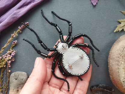 A hand holding a handmade black and white felt spider with black beads on its back, surrounded by various natural elements such as leaves, flowers, and rocks.