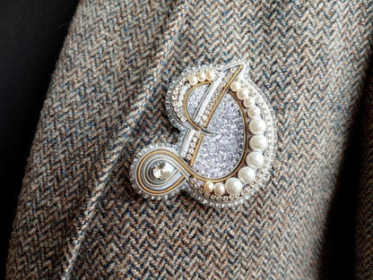A close-up image of a decorative brooch or pin with a combination of silver, white, and gold elements, including pearls and a floral design.