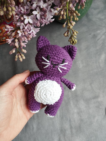 a hand holding a purple crocheted cat next to a bouquet of flowers