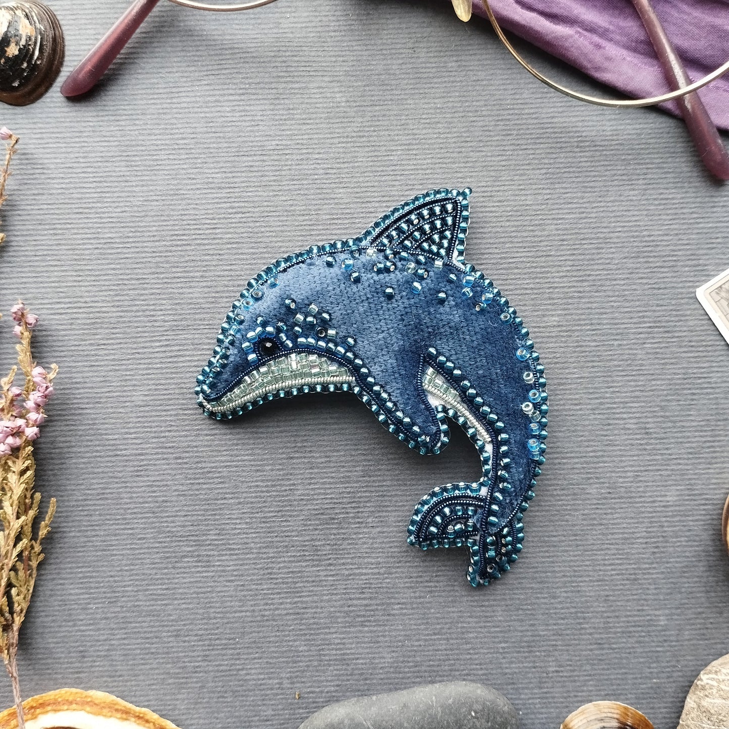 a blue, hand-made brooch or pin shaped like a dolphin, adorned with numerous small beads or sequins, resting on a grey surface.