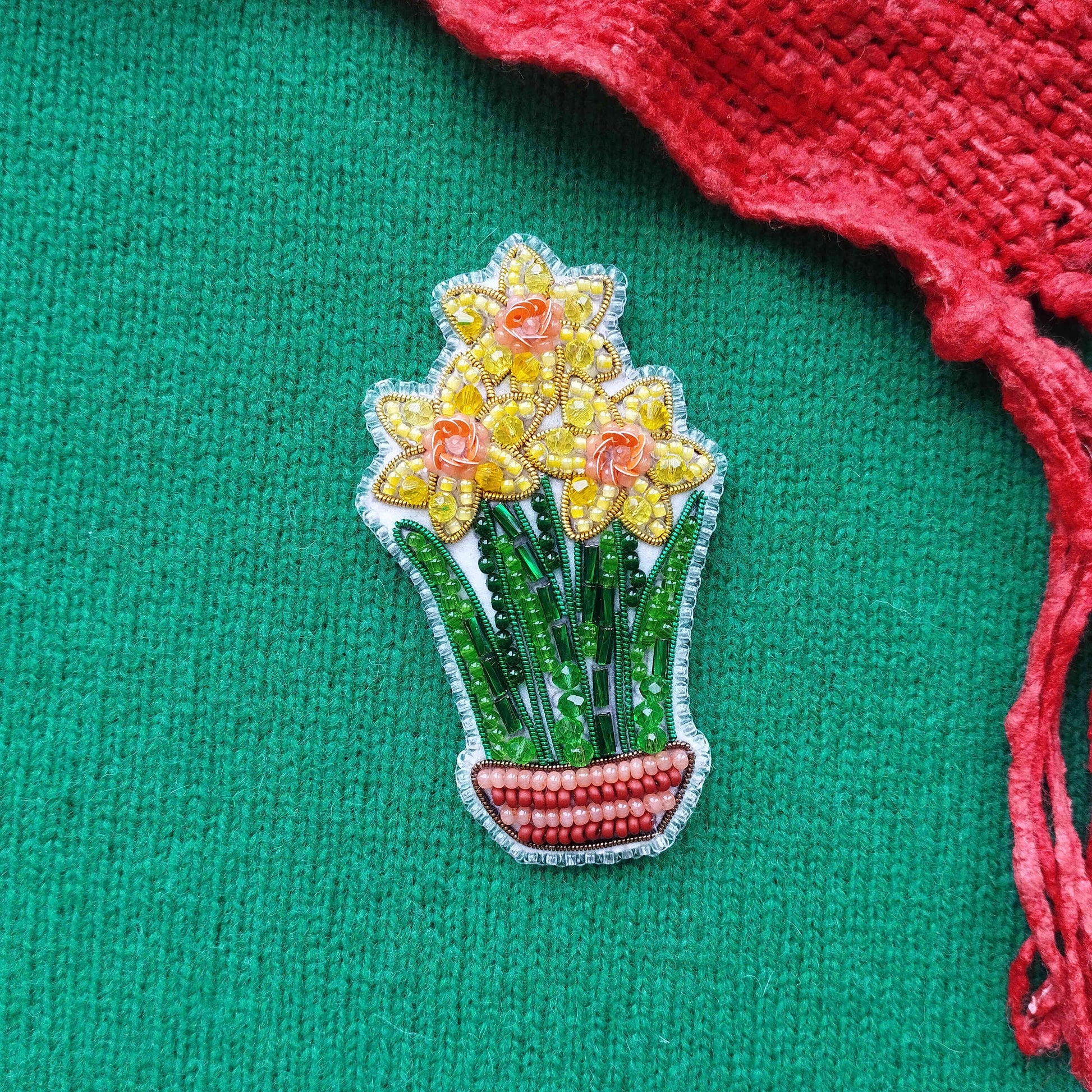 A decorative piece featuring a vase with yellow flowers and green leaves, placed on a green textured surface next to a red fabric.