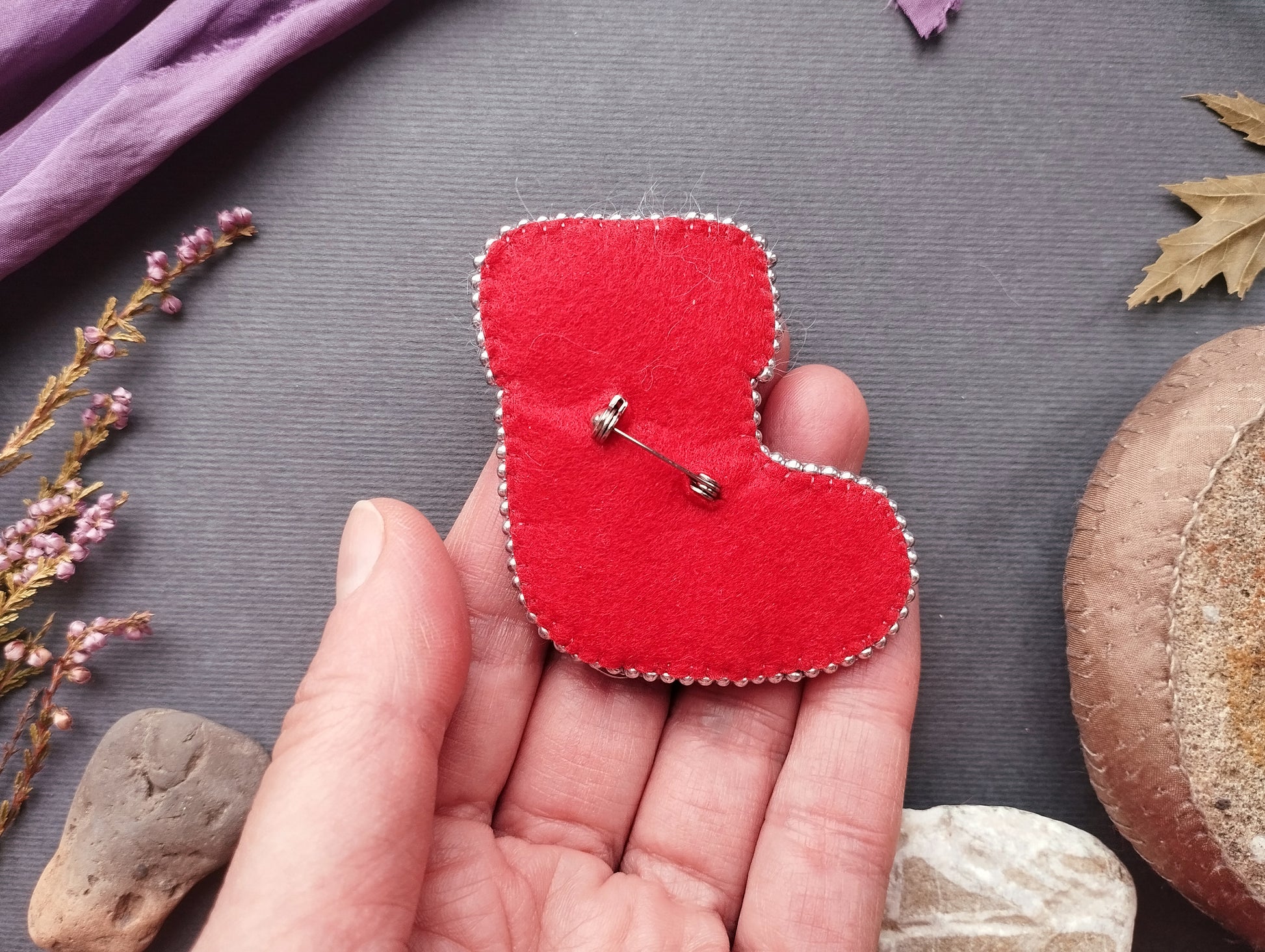 A hand holding a red felt boot pin against a gray background, surrounded by various natural elements such as leaves, flowers, and rocks.