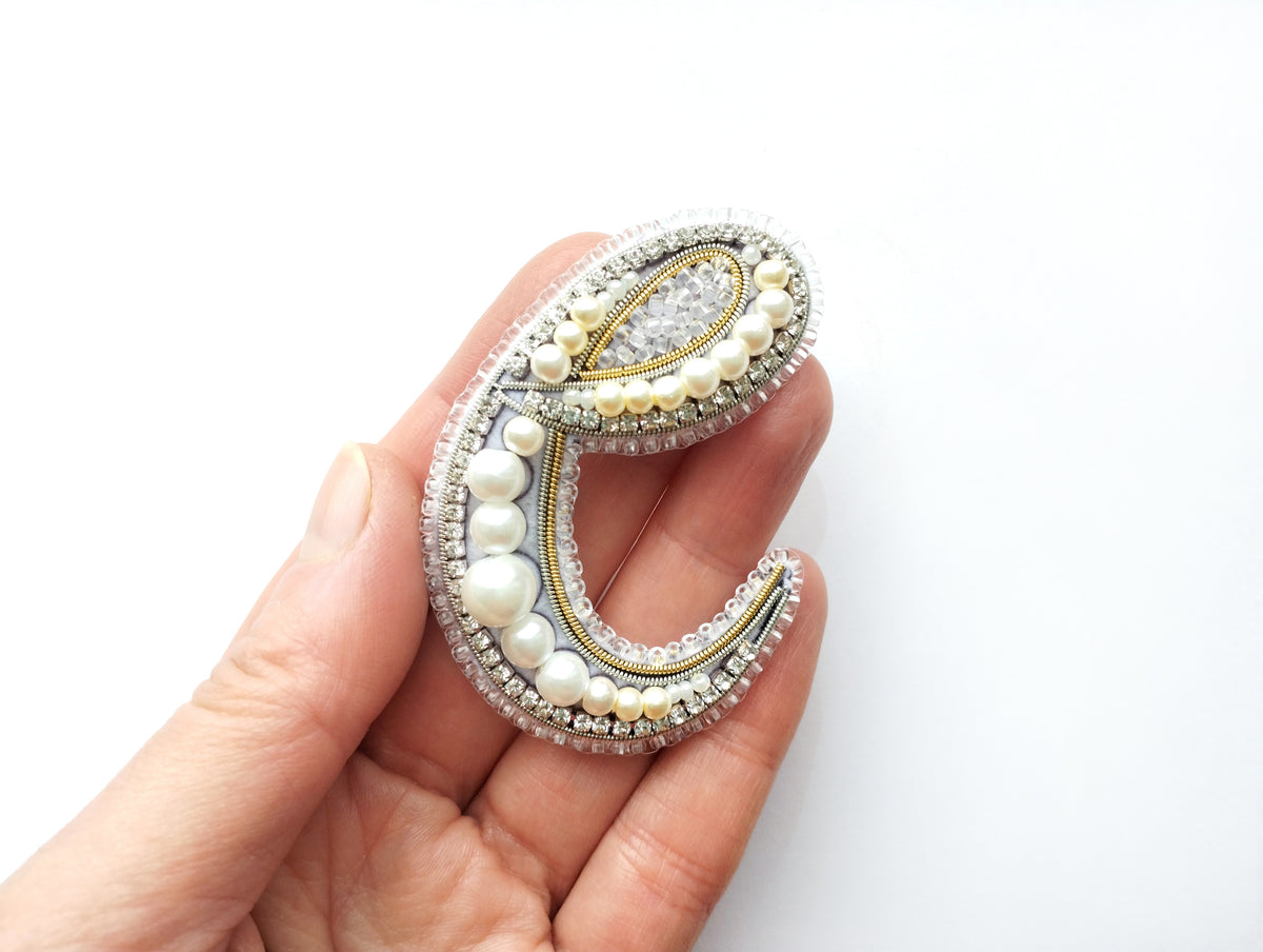 A hand holding a decorative letter 'C' made of various materials, including pearls, beads, and crystals.