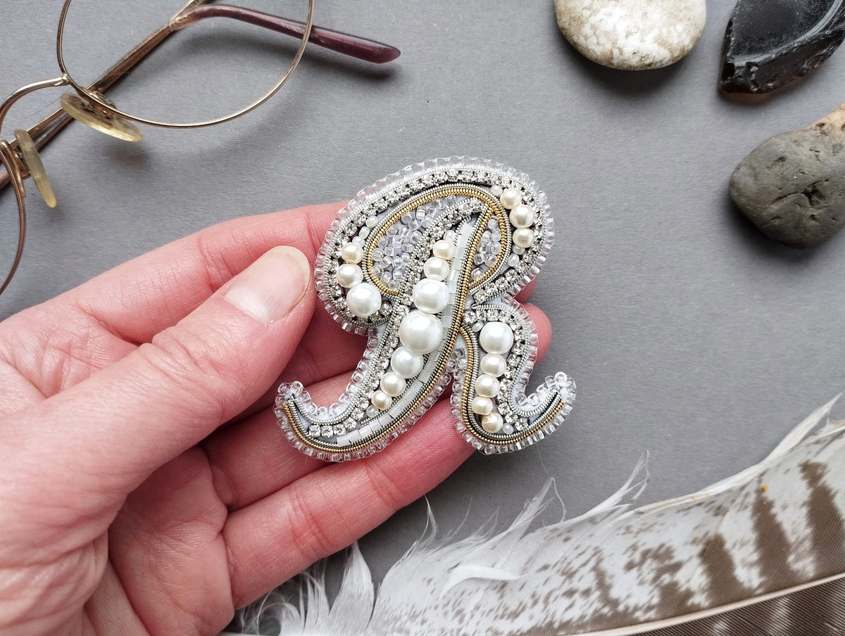 A hand holding a decorative letter "R" made of pearls and jewels, surrounded by various objects such as glasses, rocks, and feathers.