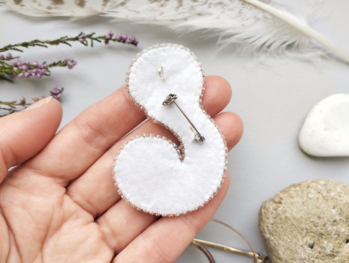 A hand holding a white felt pin with a silver pin attached to it, surrounded by various natural elements such as feathers, flowers, and rocks.
