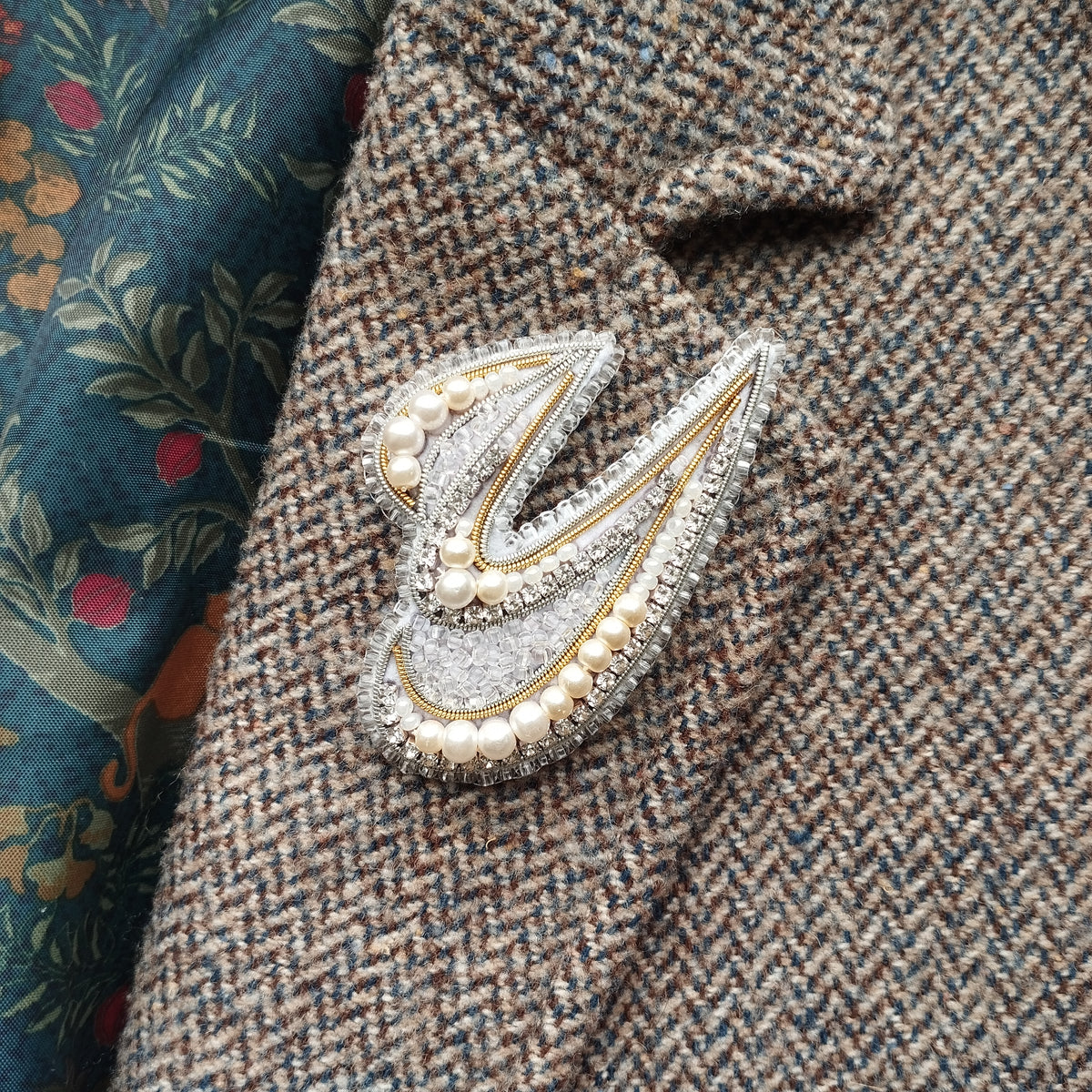 A close-up image of a decorative brooch or pin with pearls and a metallic design, attached to a tweed jacket or blazer.