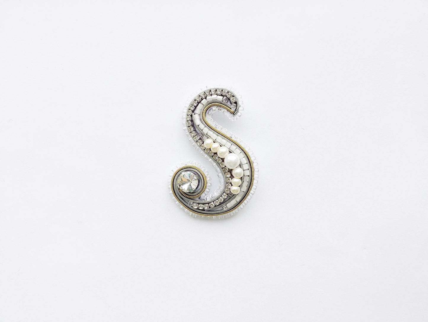 A decorative letter 'S' made of various materials, including pearls, beads, and crystals, against a plain white background.