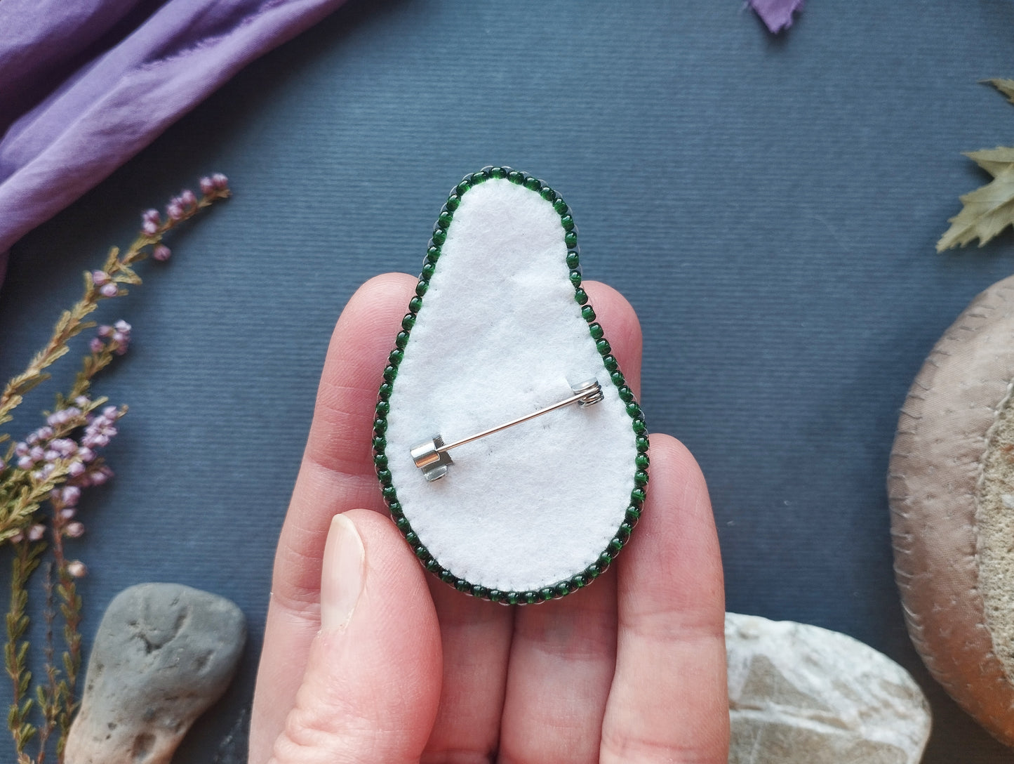 A hand holding a small, handmade pin with a green and white beaded design, placed on a blue surface surrounded by various natural elements such as flowers and rocks.