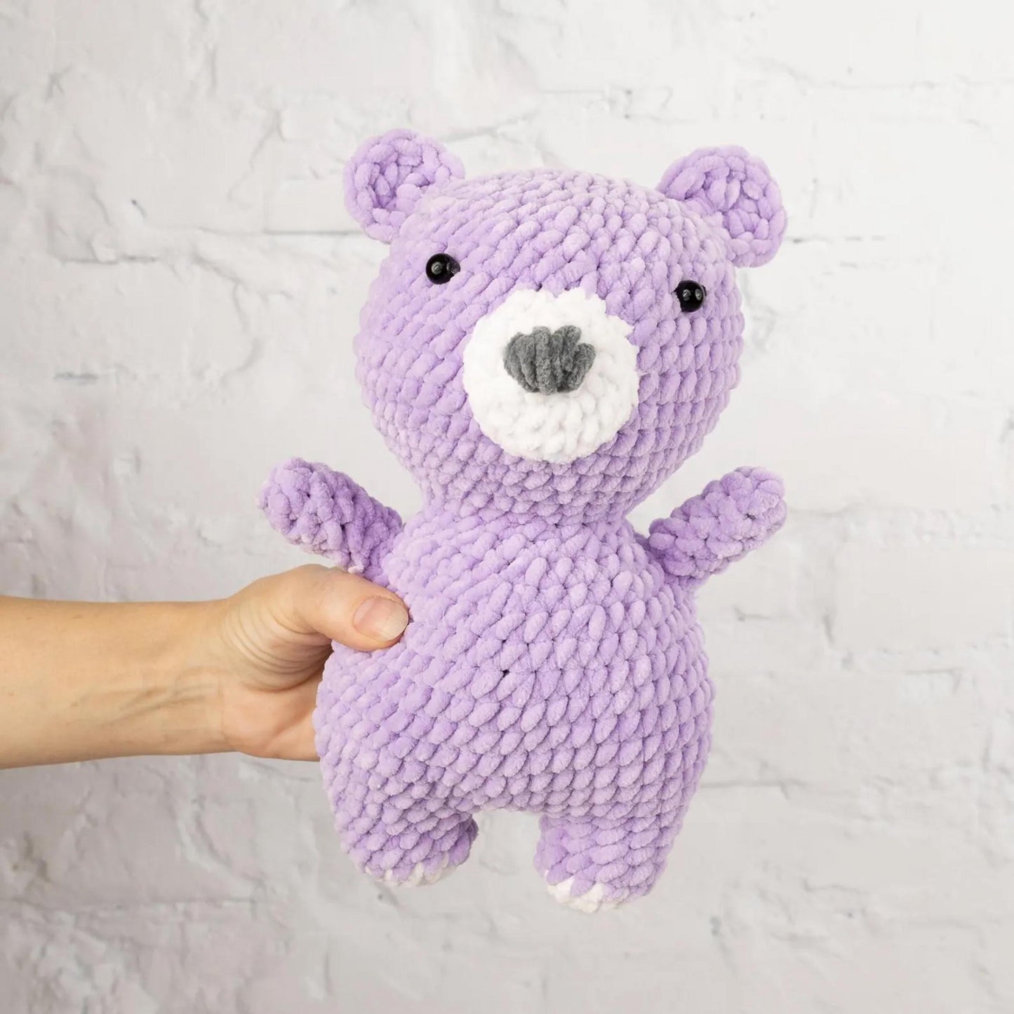 A hand is holding a purple crocheted teddy bear against a white brick wall background.