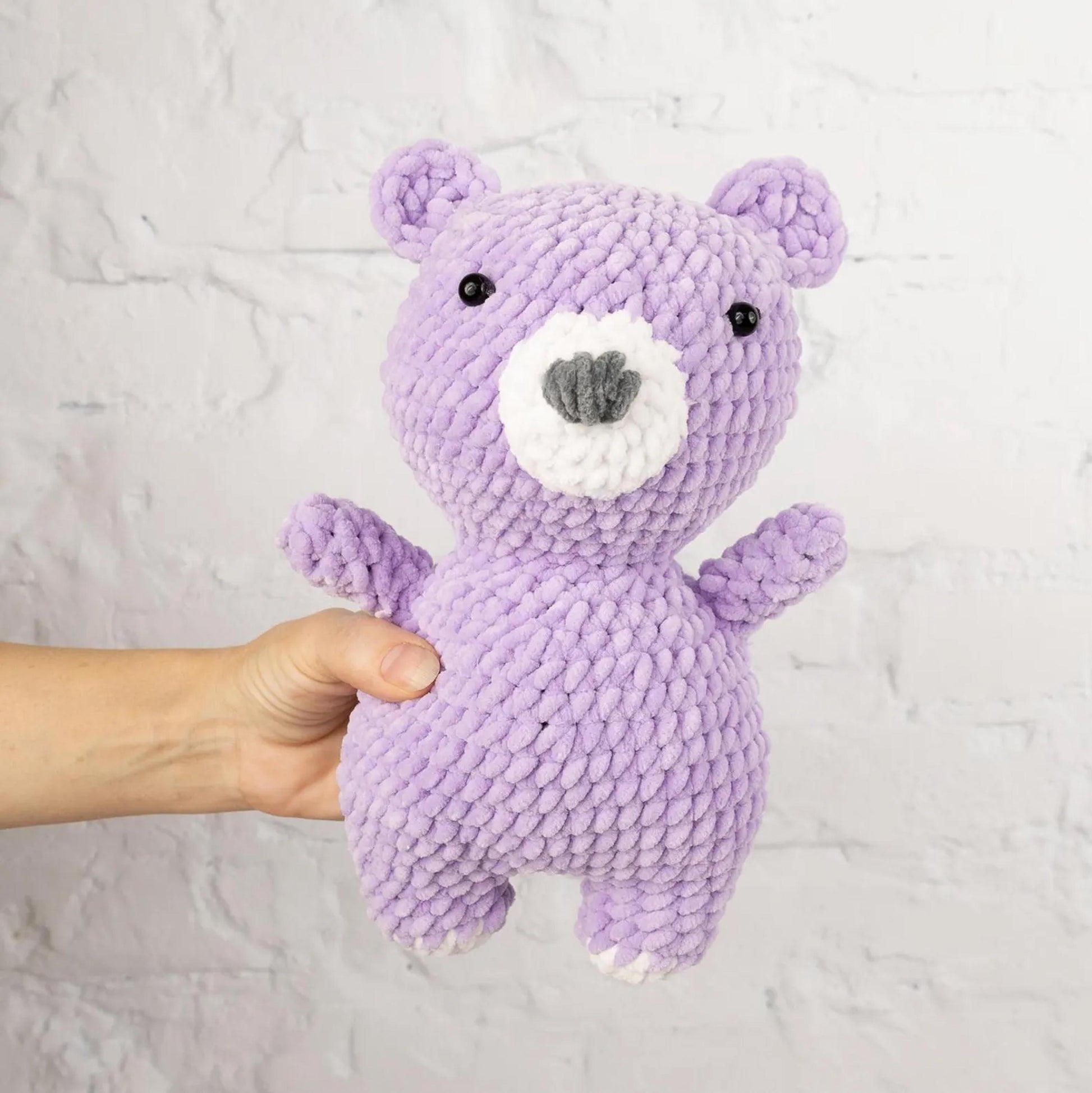 A hand is holding a purple crocheted teddy bear against a white brick wall background.