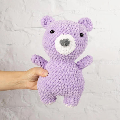 A hand is holding a purple crocheted teddy bear against a white brick wall background.