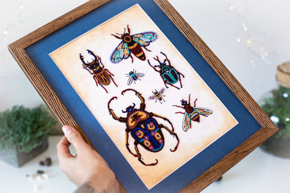 A hand is holding a framed picture of various colorful insects, including a large beetle and several smaller insects, against a blue background.